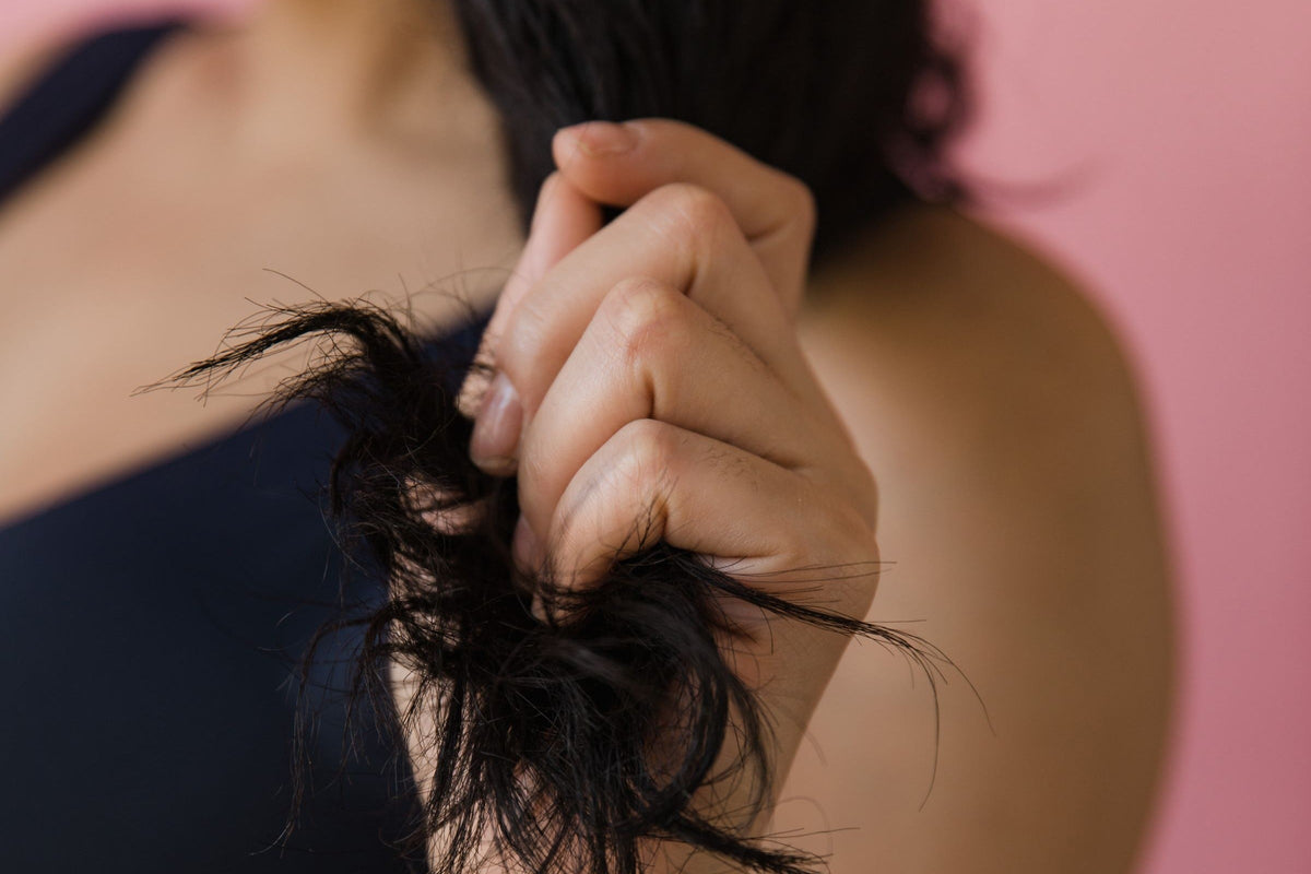 Main tenant une mèche de cheveux foncés ondulés dont les pointes sont cassées et emmêlées, sur fond rose