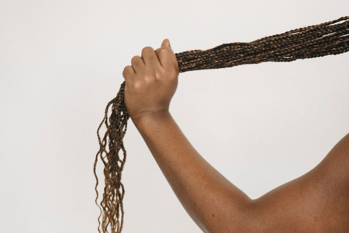 Main tenant une longue natte de box braids brunes devant un mur blanc