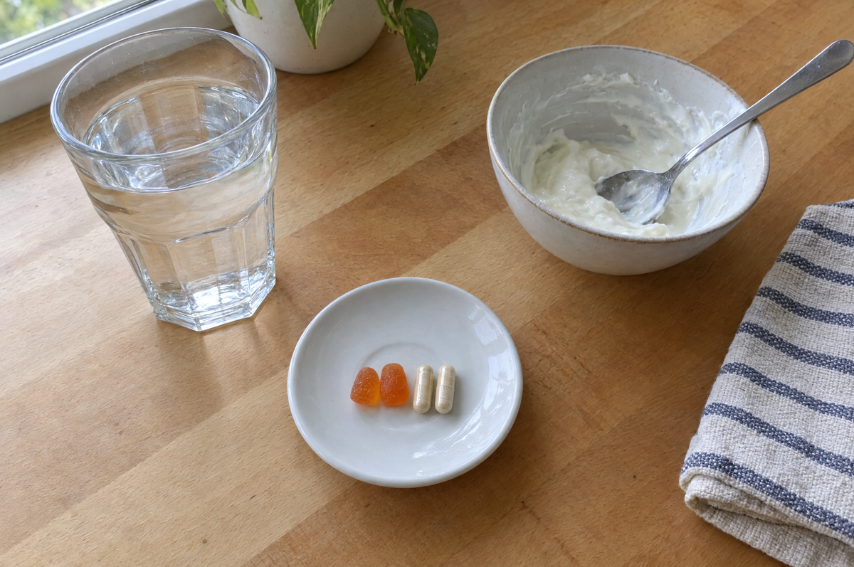 Deux gummies orangés et deux gélules posés côte à côte dans une petite coupelle blanche, sur un plan de travail en bois, à côté d'un verre d'eau et d'un bol de yaourt entamé