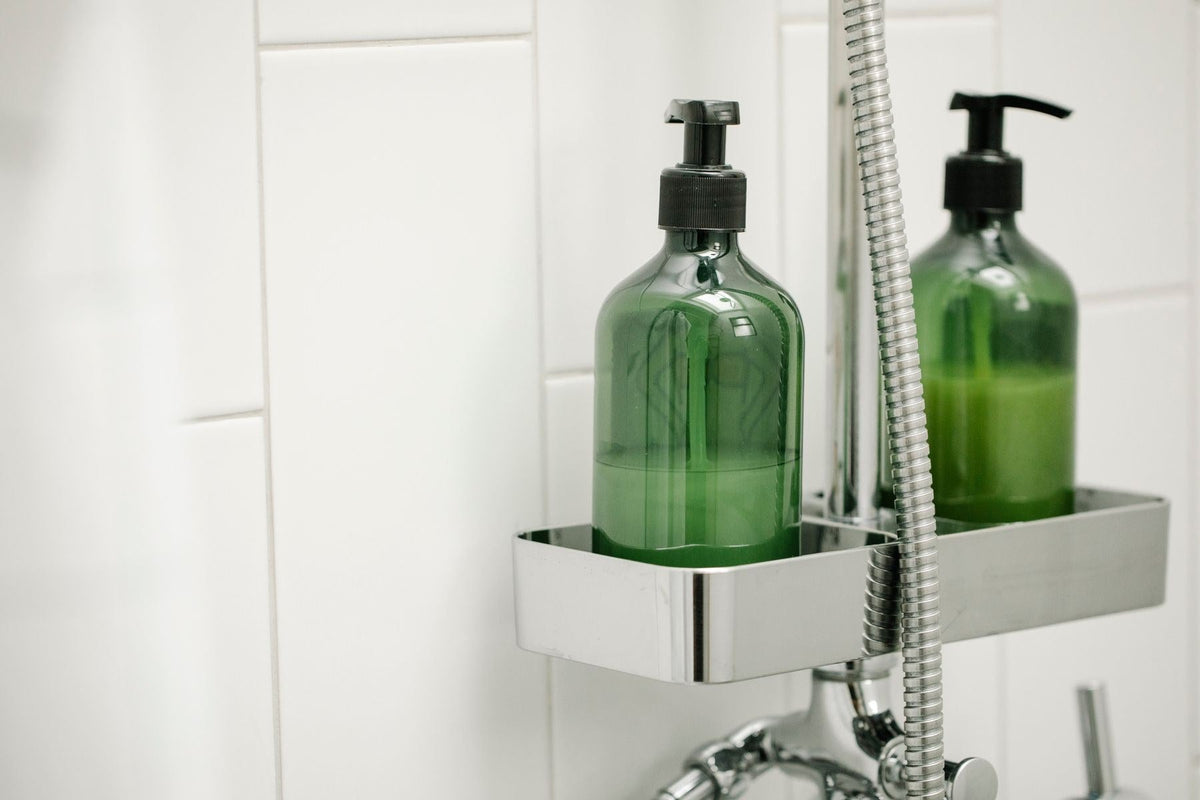 Deux flacons pompe verts posés sur une étagère de douche chromée, contre un mur de carrelage blanc